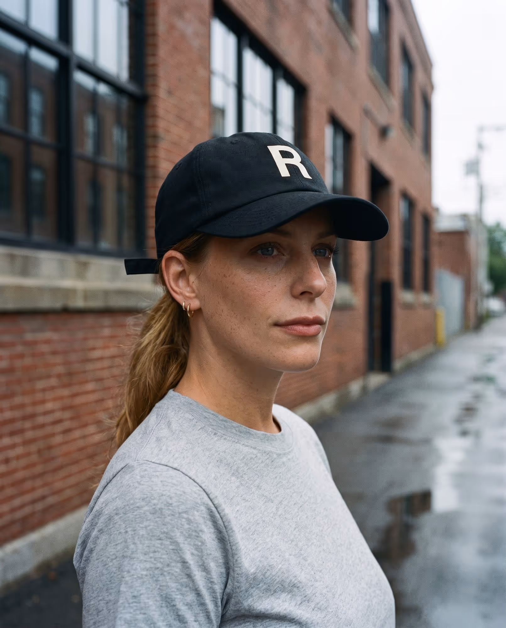 Model wearing a black structured cap with a single printed cream letter on the front, photographed on a Cambridge street