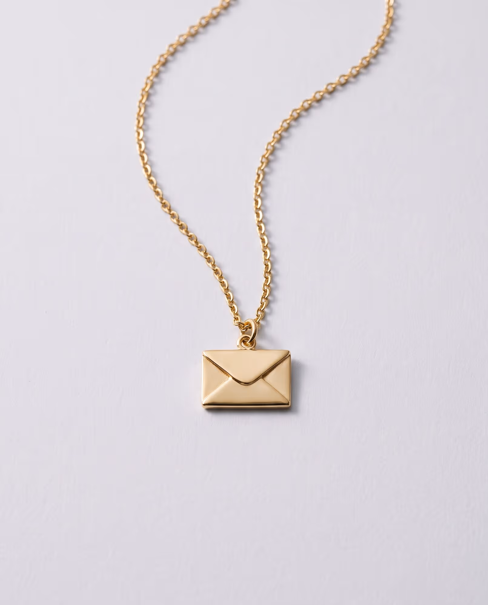 Blank gold-tone envelope pendant on a fine chain, laid flat on a pale grey backdrop