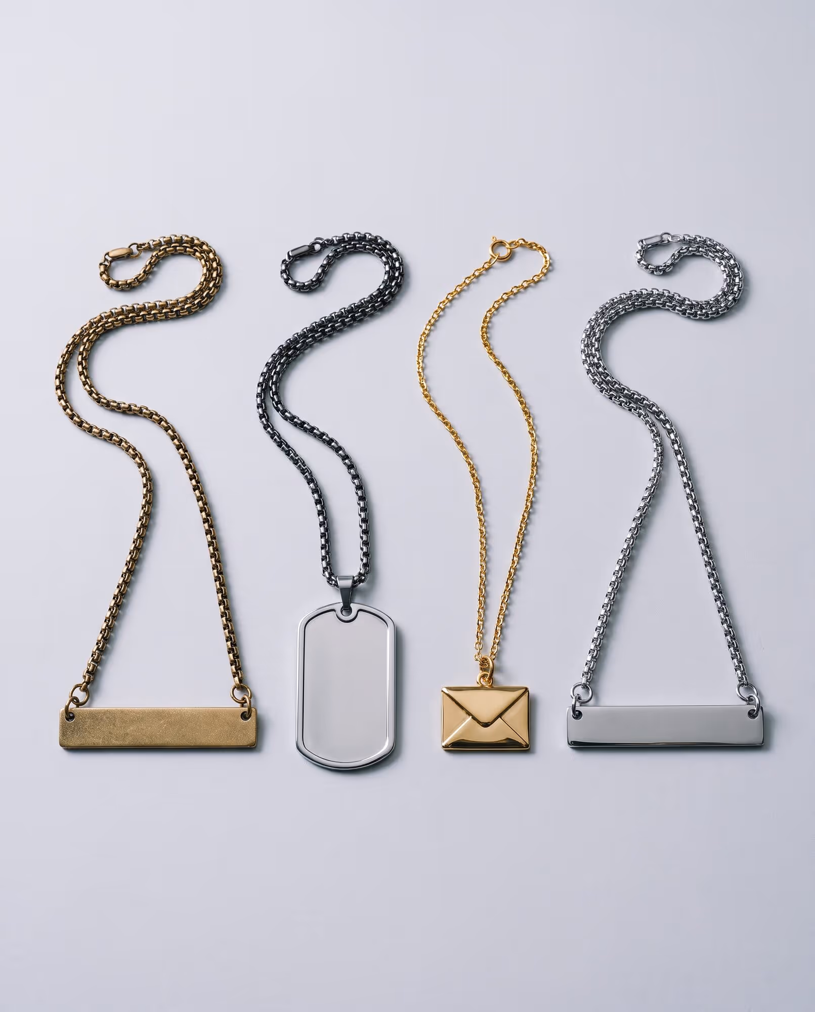Four blank necklaces laid flat on a pale grey backdrop — a gold-tone bar, a silver-tone dog tag, a gold-tone envelope pendant and a silver-tone bar