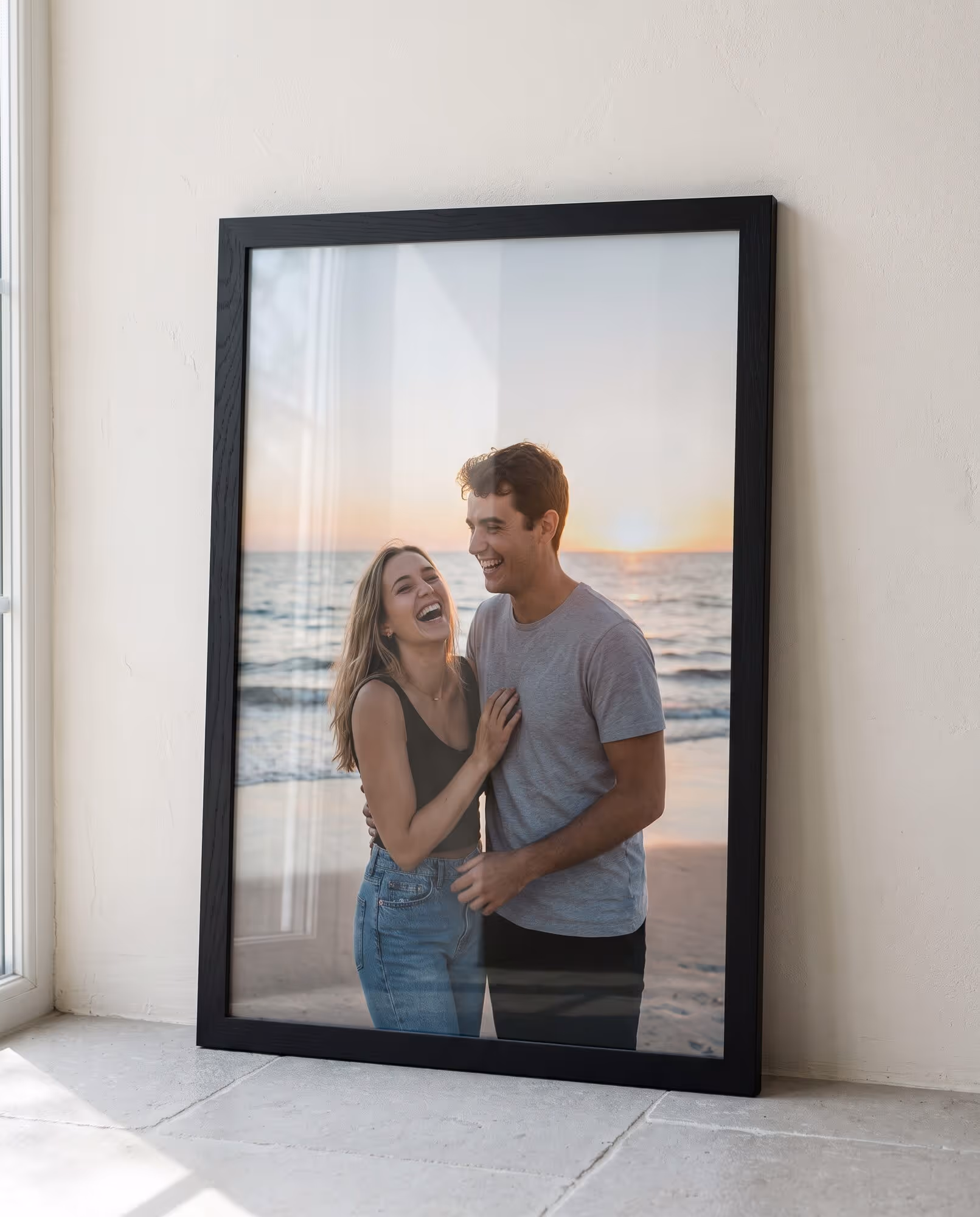 Black wooden portrait frame holding a beach photo of a laughing couple, leaning against a pale wall on a tiled floor