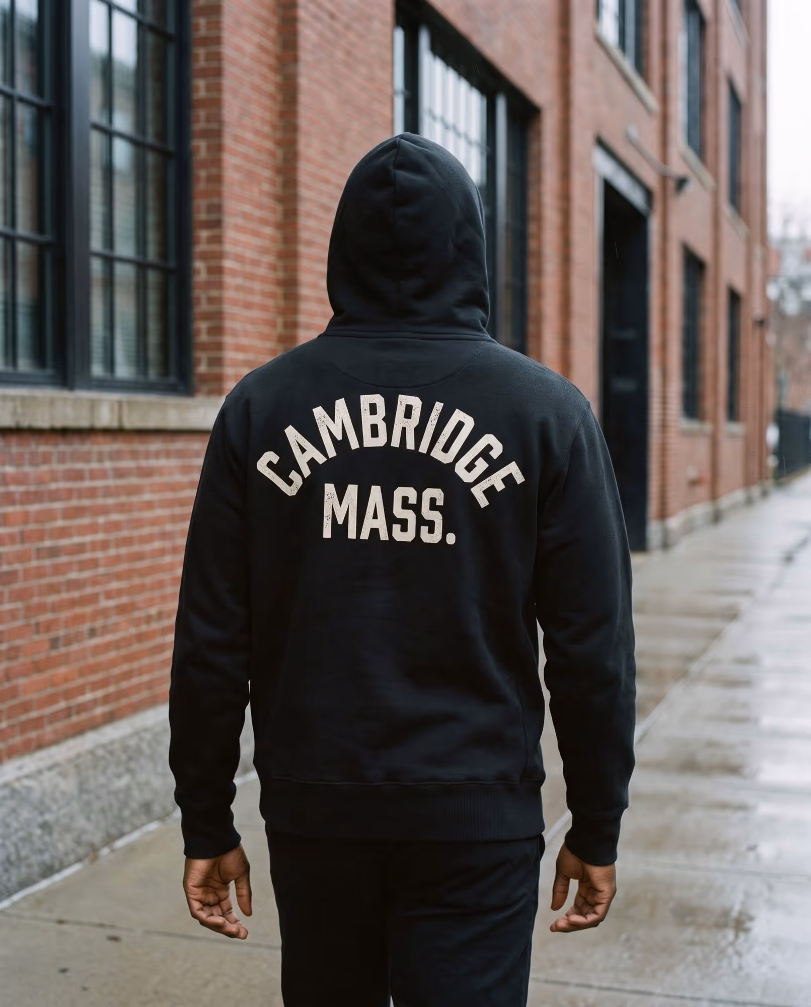 Back of a black pullover hoodie with "Cambridge Mass." printed in cream collegiate lettering, hood up