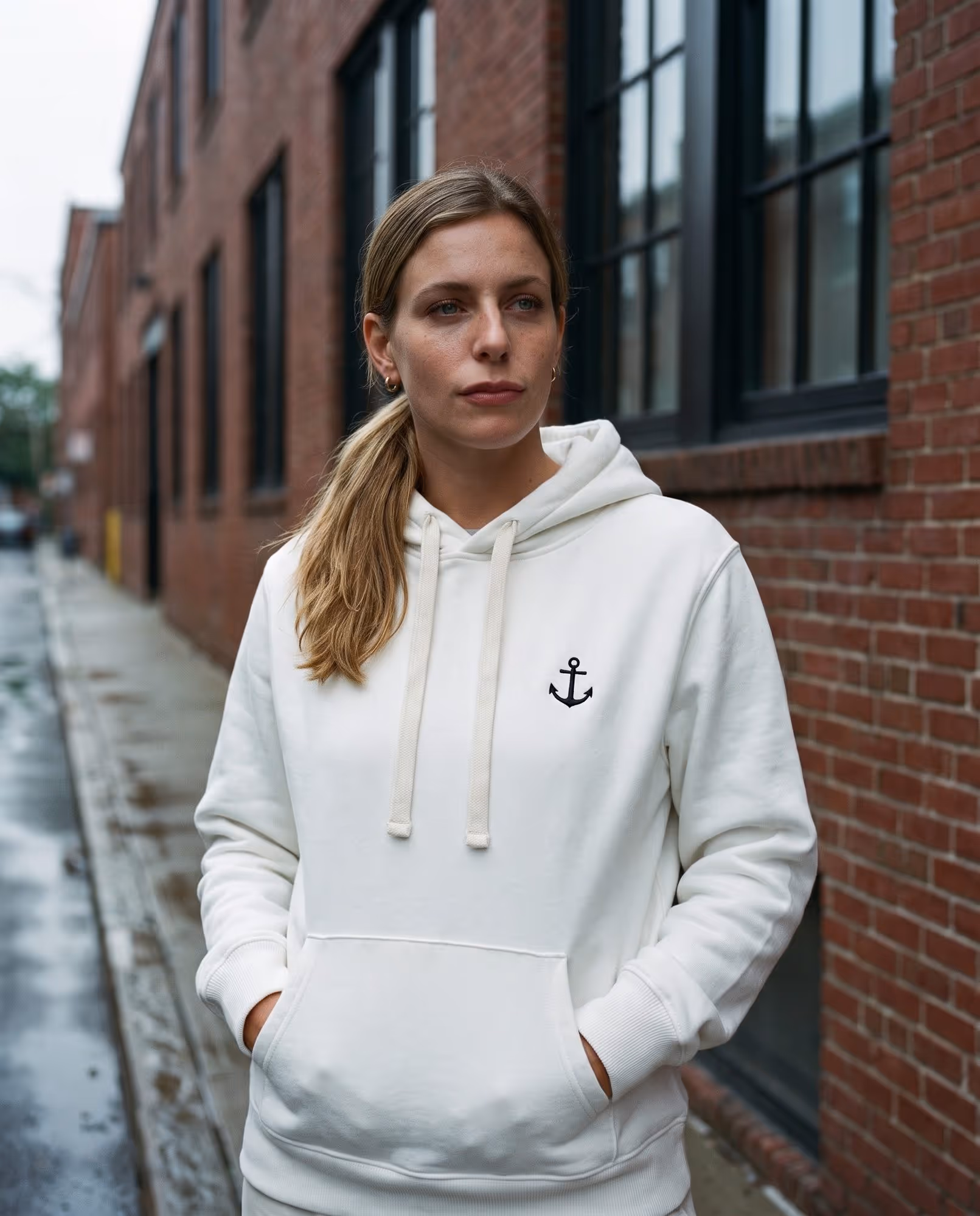 Model wearing a white pullover hoodie with a small navy anchor printed on the chest, photographed on a Cambridge street
