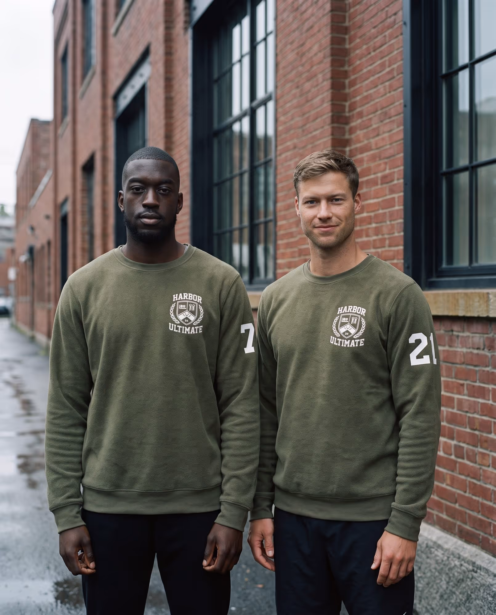 Two models in matching olive crewneck sweatshirts with a team crest and sleeve numbers