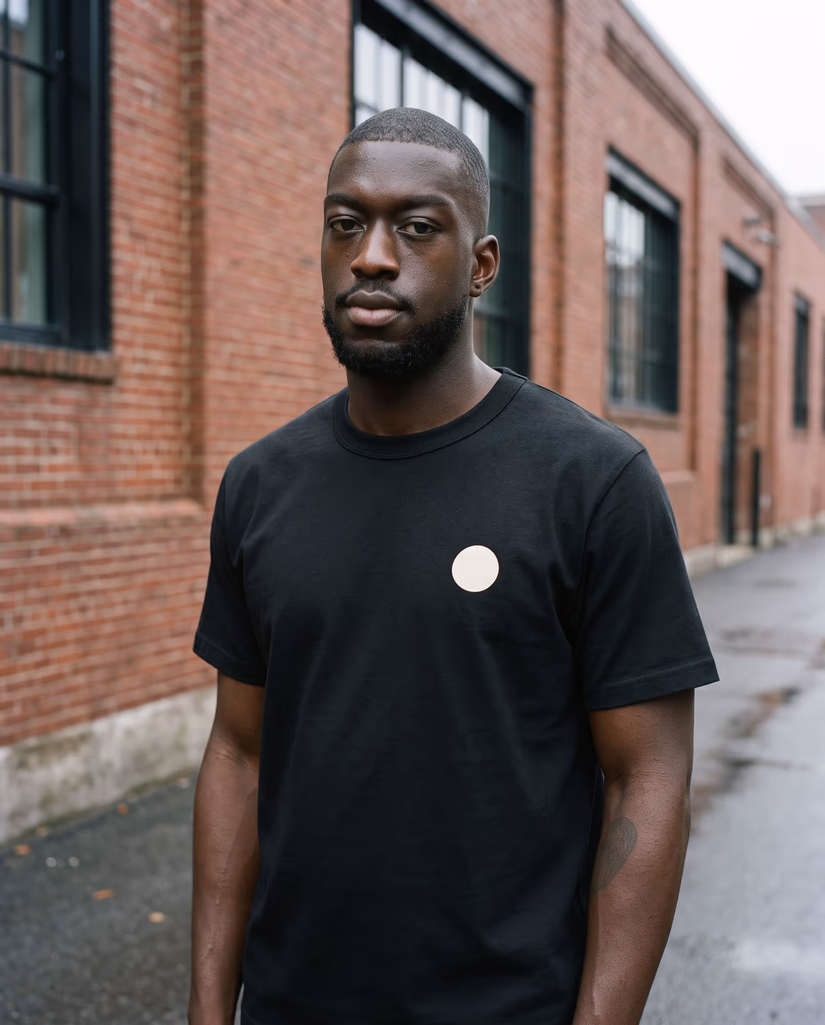 Model wearing a black custom-printed heavyweight t-shirt with a small cream chest mark, photographed on a Cambridge street