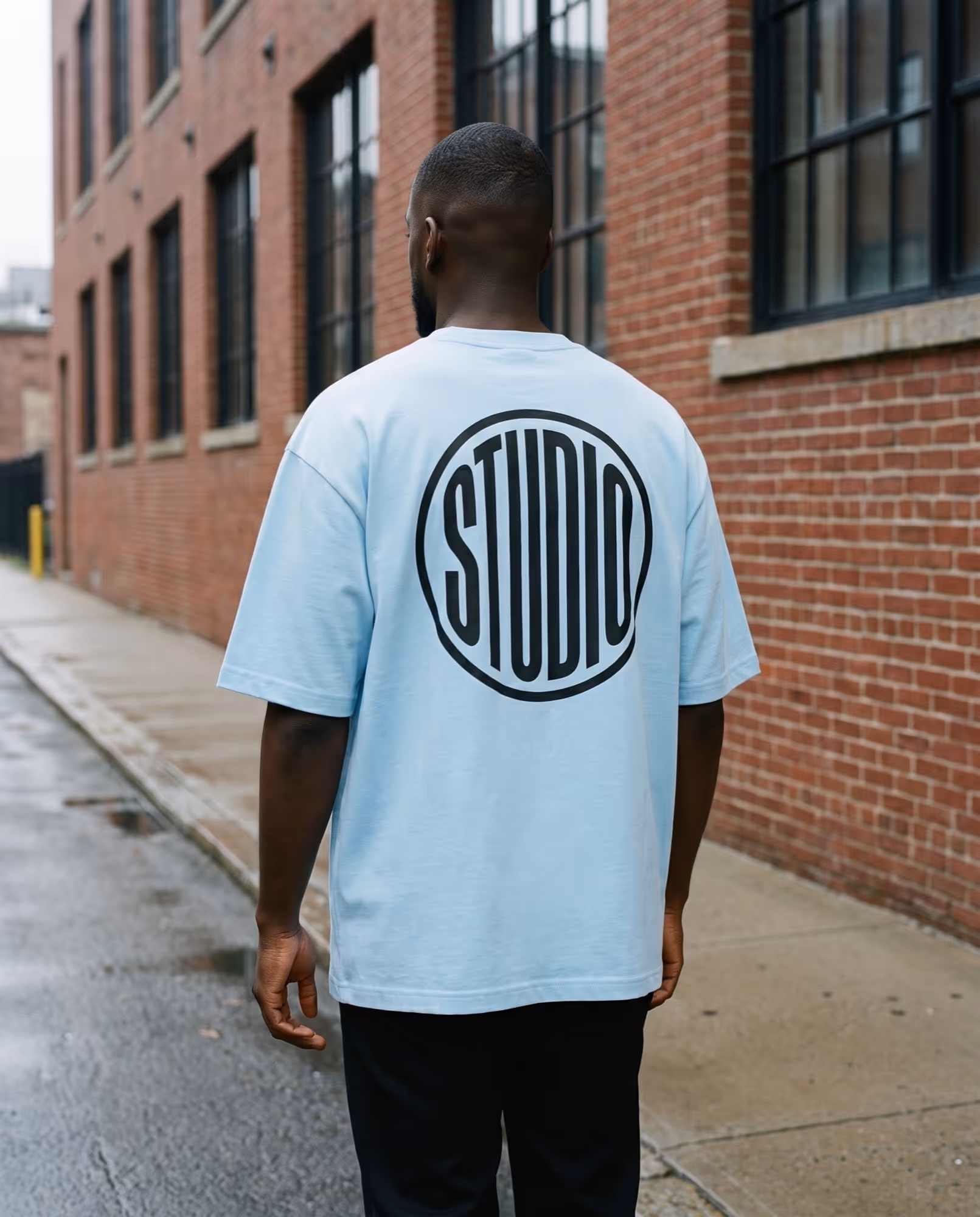 Sky blue oversized t-shirt with a large black circular STUDIO emblem across the upper back
