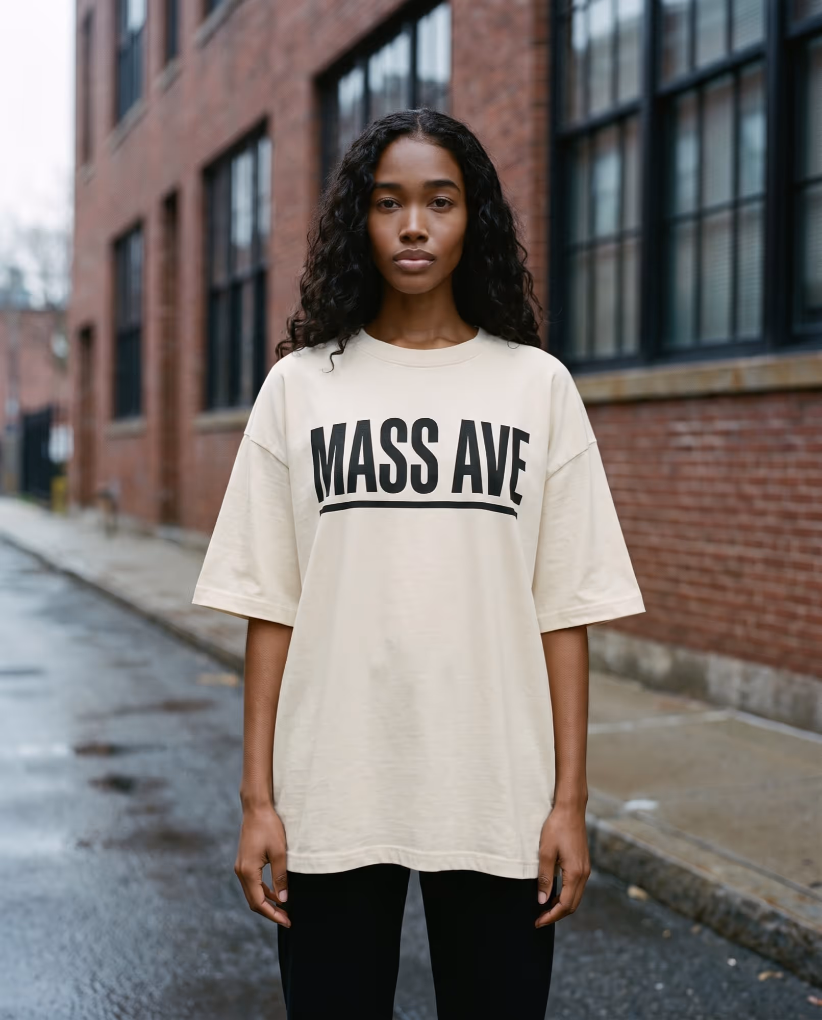 Model wearing a cream oversized boxy t-shirt printed with MASS AVE in black, photographed on a Cambridge street