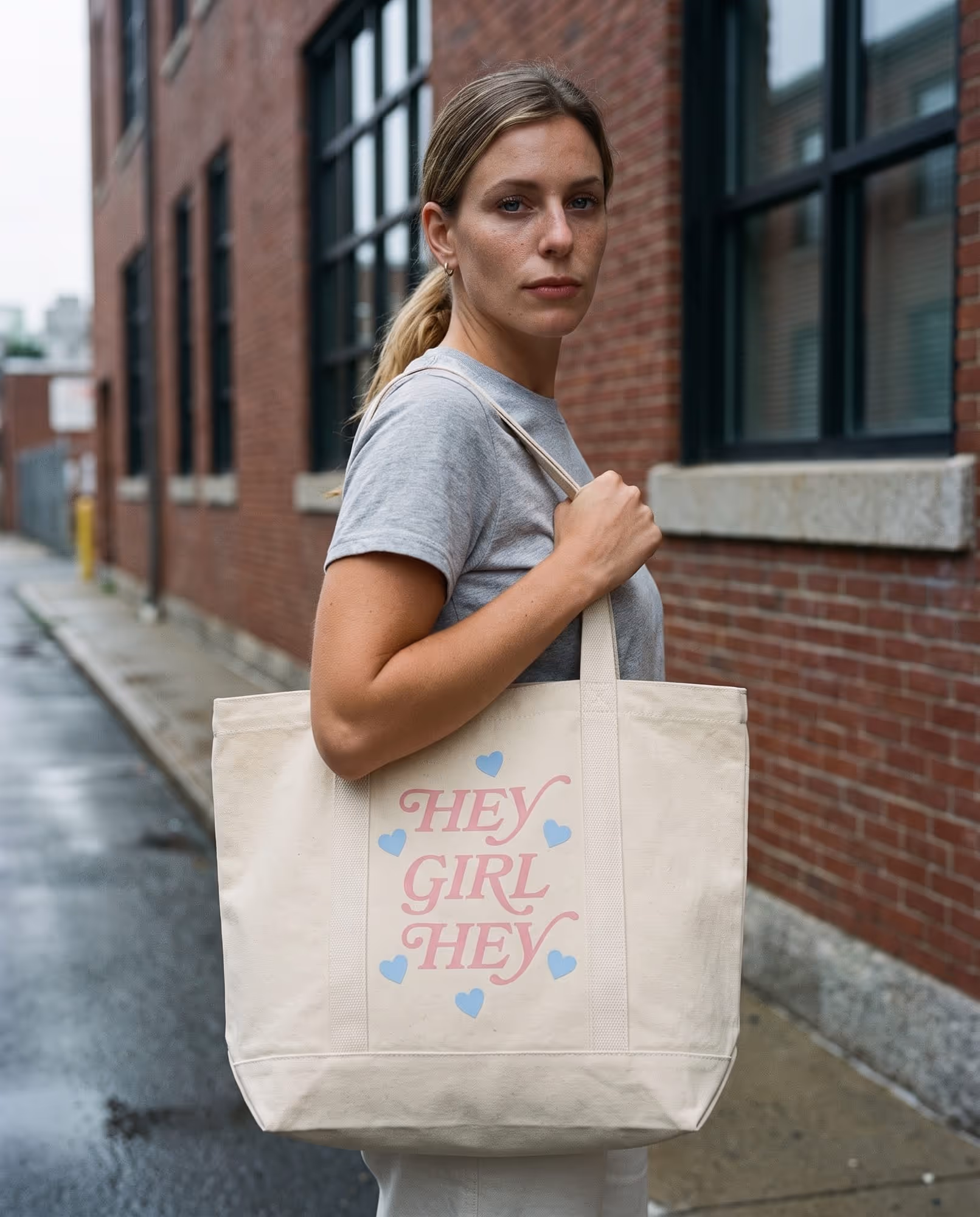 Natural canvas tote bag with "HEY GIRL HEY" printed in pink script surrounded by small blue hearts