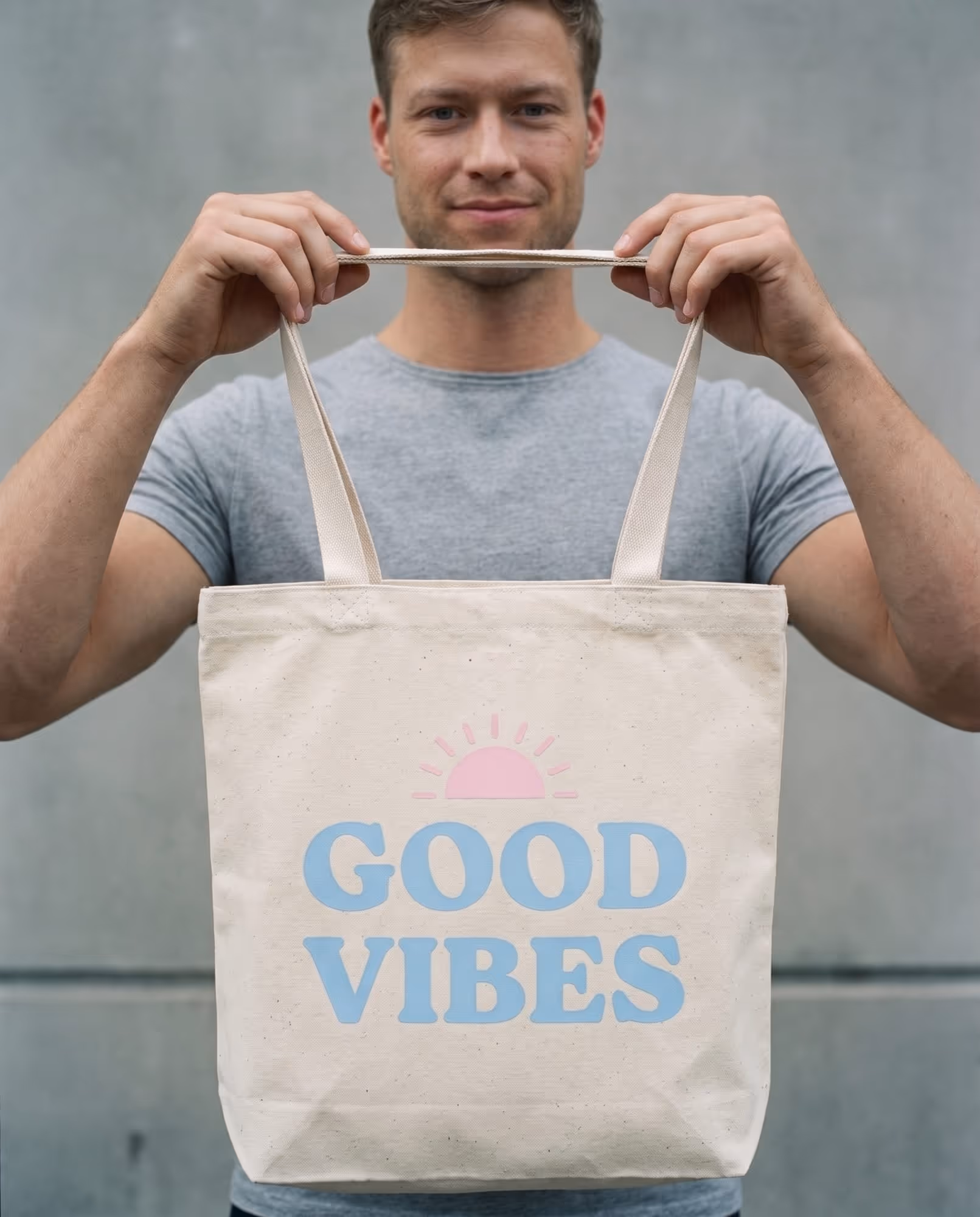 Model holding up a natural canvas tote bag printed with "GOOD VIBES" in pale blue rounded capitals under a small pink sun